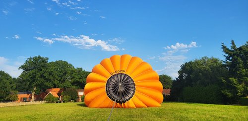 Der orangefarbene Heißluftballon füllt sich. Er liegt noch auf der Wiese, sodass die spätere Oberseite mit der dunklen Abdeckung zu sehen ist.