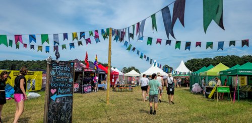 Im Vordergrund sind Menschen zu sehen, die an einem Festivalstand entlanggehen, dekoriert mit bunten Wimpeln und Bannern.
