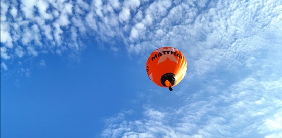 Der orangefarbene Heißluftballon schwebt in der Luft, der Himmel strahlt blau, mit kleinen weißen Wolken