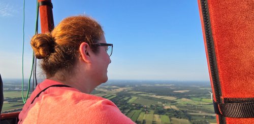 Verena schaut vom Heißluftballon aus in die Weite, rechts ist eine rot ummantelte Stande zu sehen