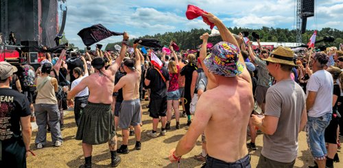 Festivalbesucher, viele ohne Oberbekleidung, feiern und schwenken ihre Shirts vor der beeindruckenden Bühnenkulisse.