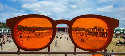 Sonnenbrille im Vordergrund, die ein lebhaftes Festivalgelände mit Zuschauern und einem Bühnenaufbau zeigt.