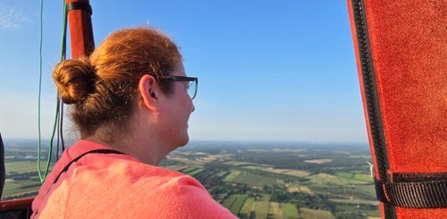 Verena schaut vom Heißluftballon aus in die Weite, rechts ist eine rot ummantelte Stande zu sehen