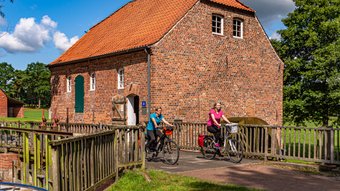 Zwei junge Radlerinnen fahren über die Brücke vor einer hitsorischen Wassermühle aus Backstein