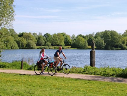 Ein Paar radelt im Frühling am Vörder See entlang, im See ist das Kunstwerk Seemann zu sehen