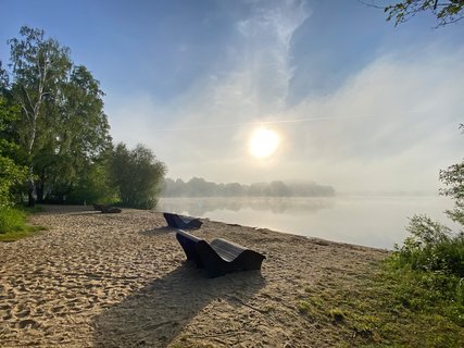 Relaxliegen aus Holz stehen am Strand des Vörder Sees, der von Morgennebel bedeckt ist